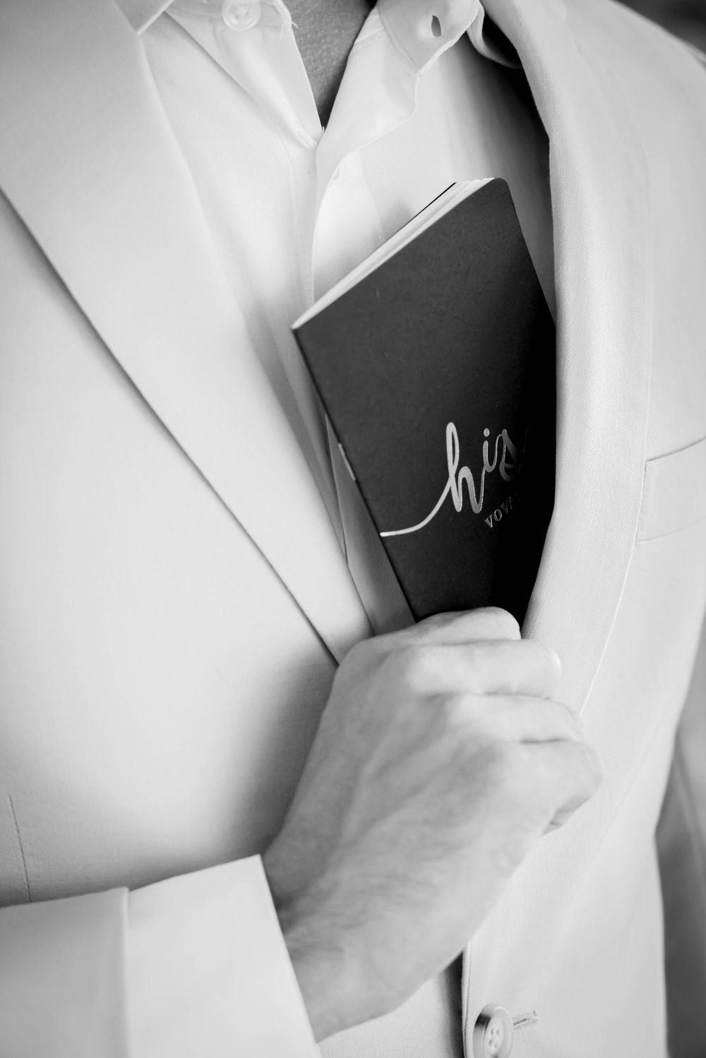 A black vows book being held inside a white suit jacket pocket in black and white.