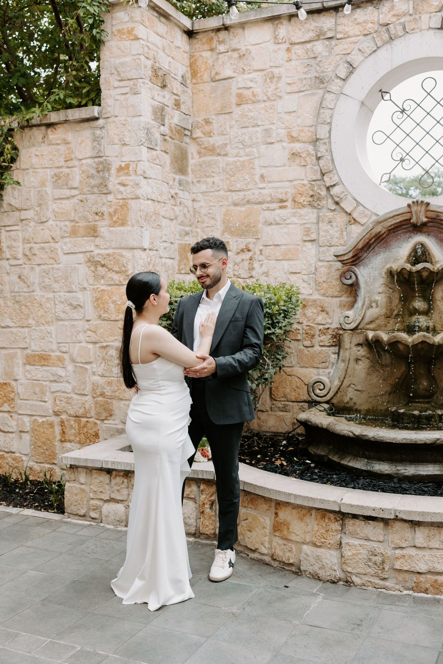 San Antonio Wedding Photography. Elegant couple against stone wall with ornate fountain and circular window in romantic outdoor setting.