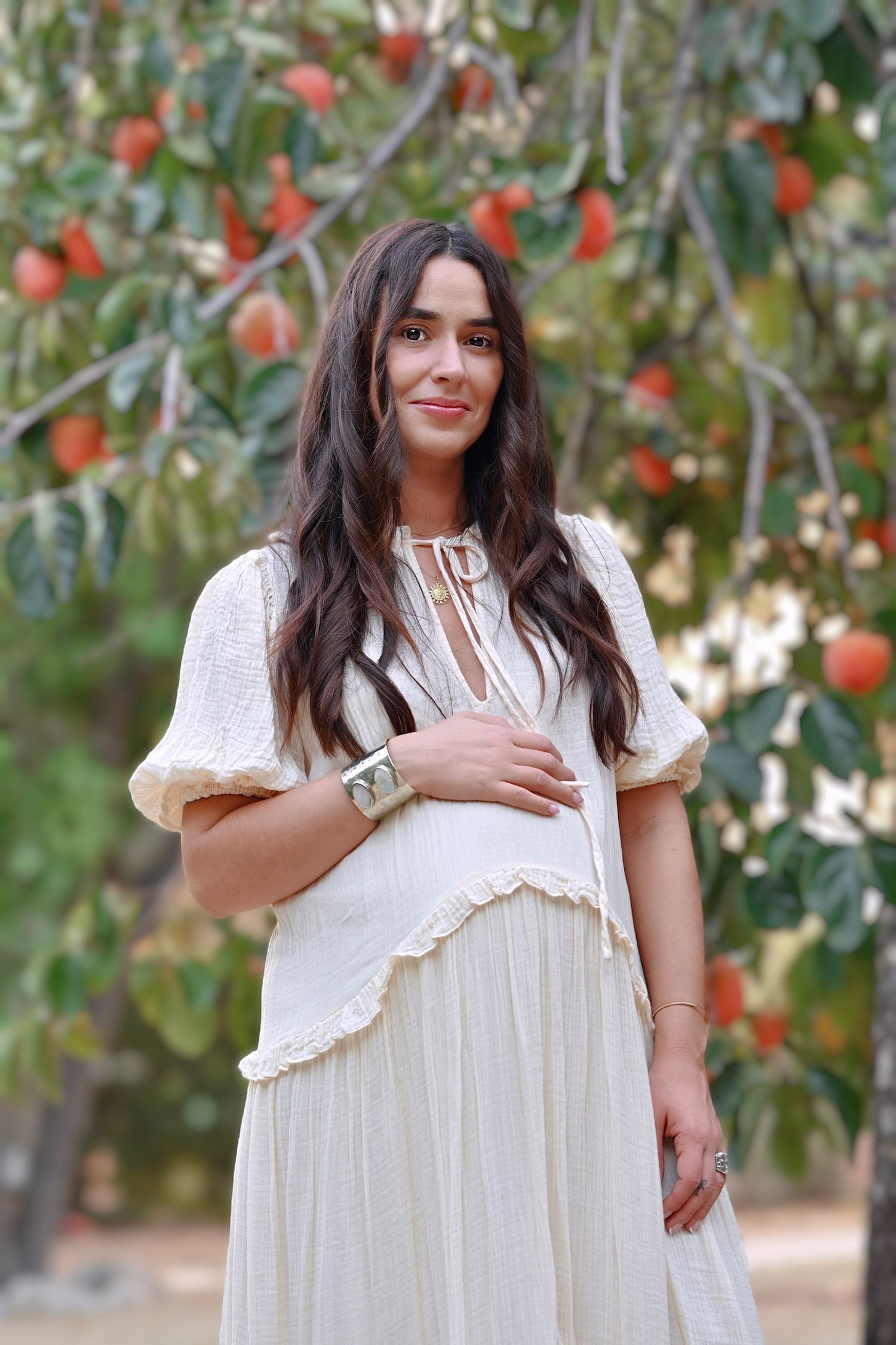 Expectant mother in flowing white dress poses by orange fruit trees in garden setting.