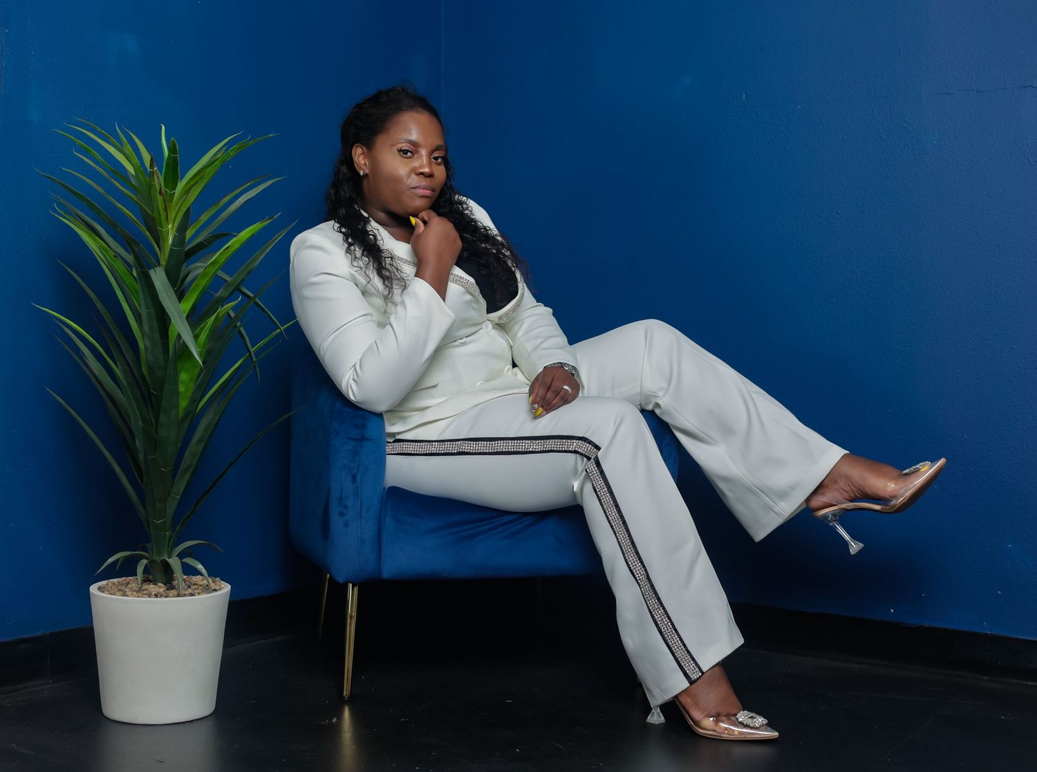 Stylish woman in white pantsuit seated in blue velvet chair beside indoor plant in blue room.