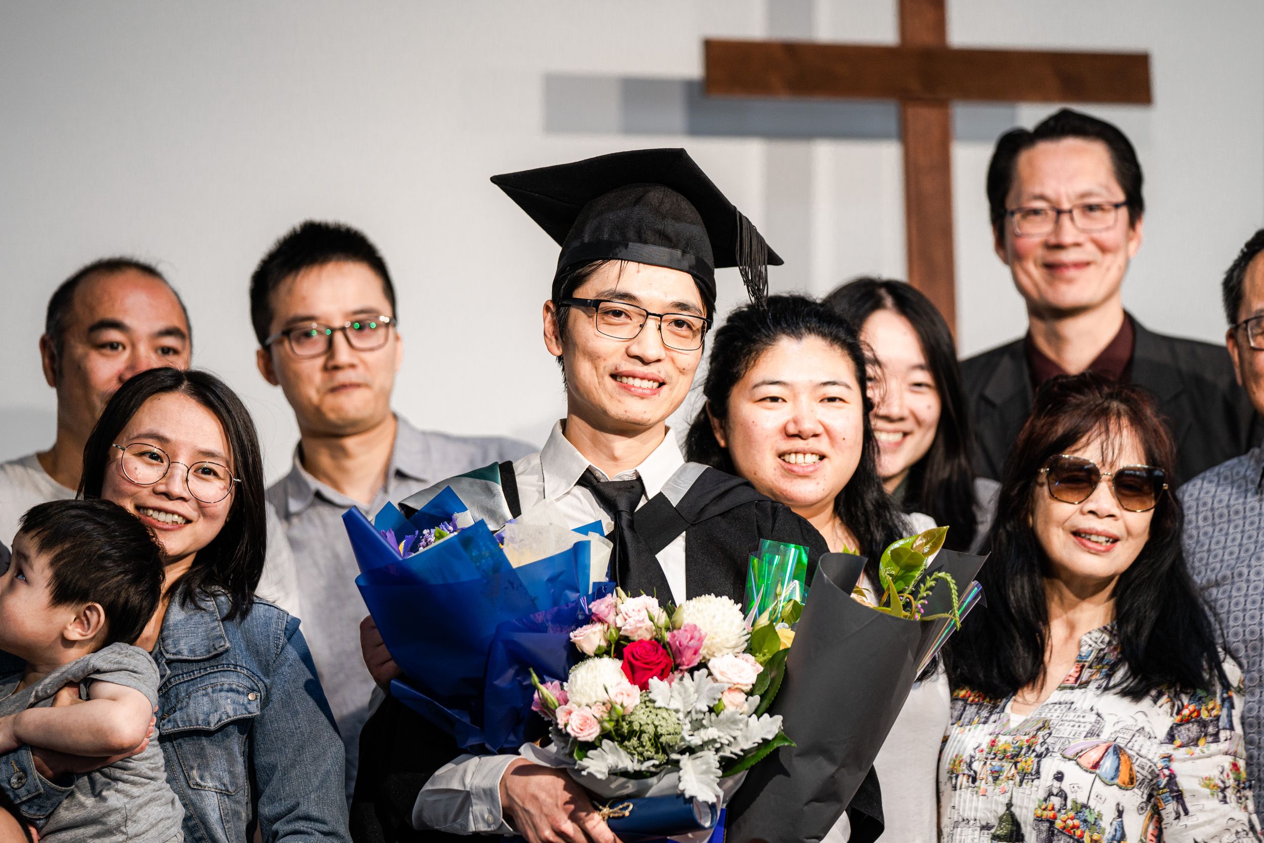 A graduation celebration with family members posing together with flowers and a cross visible in the background.