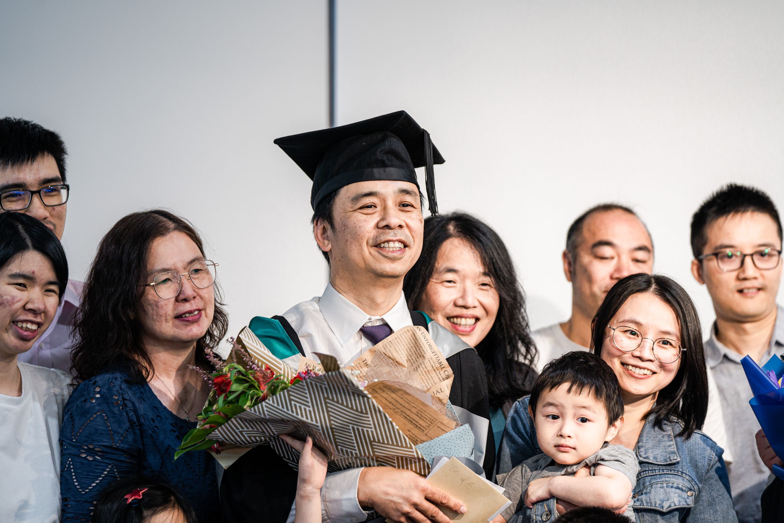 Graduate celebrating with family members and holding bouquet against white background.