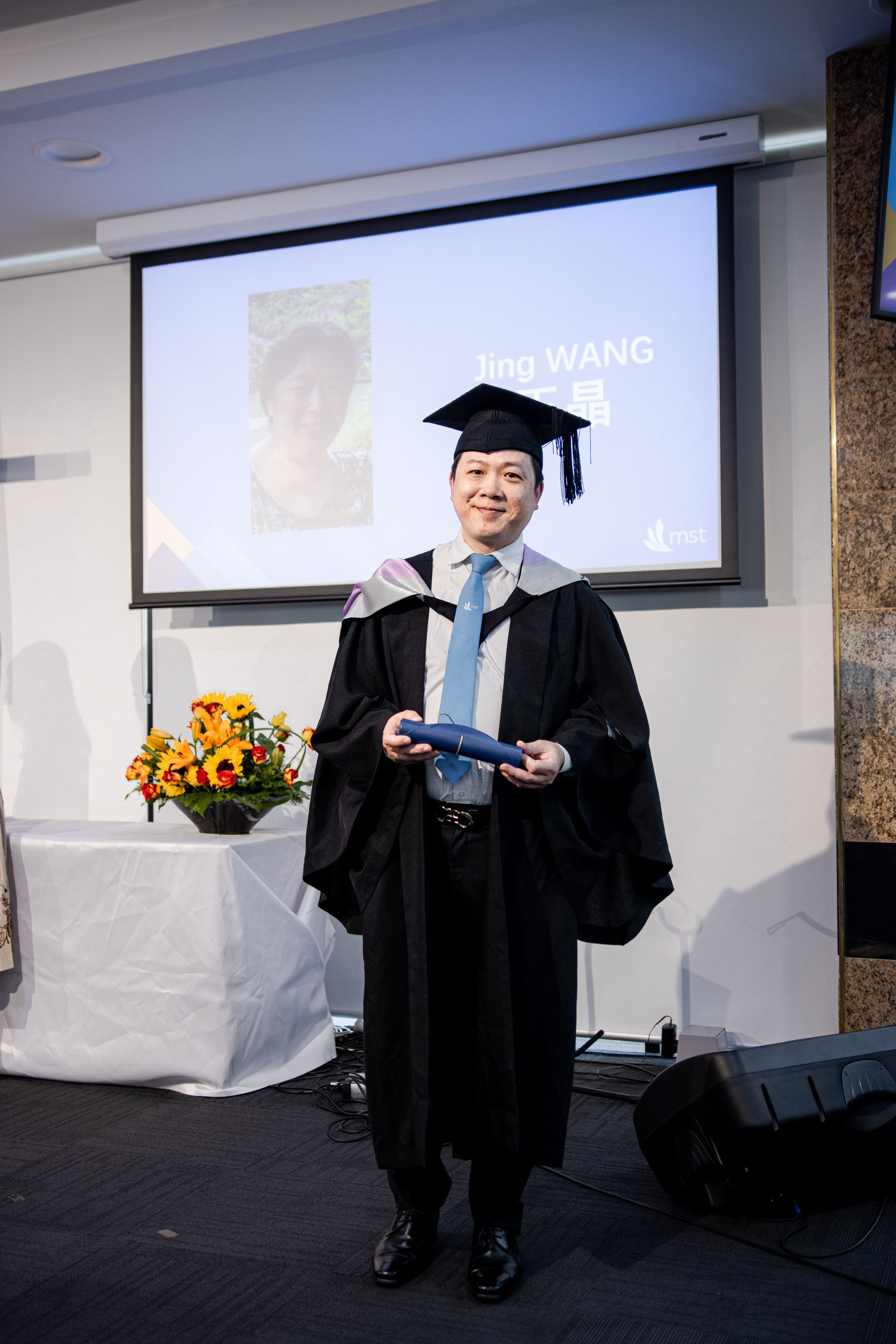 Graduate in academic regalia standing in front of a presentation screen with flowers.
