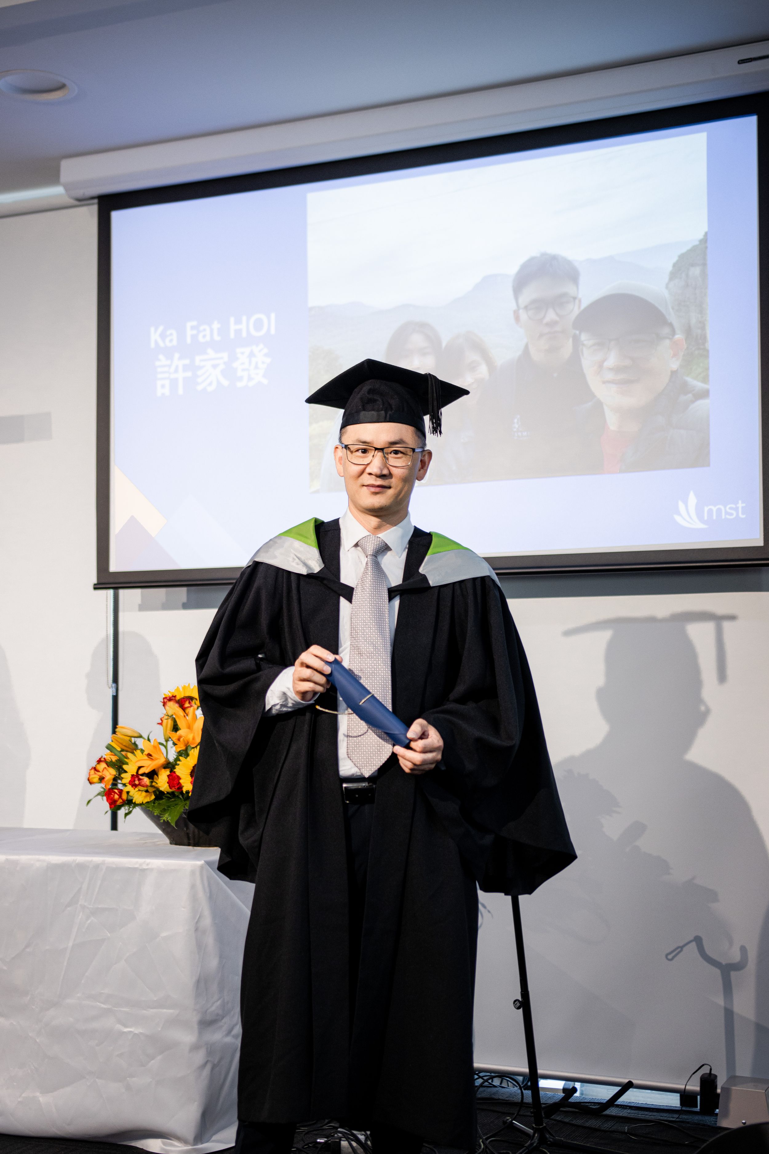 Graduate posing with diploma in front of projection screen with floral arrangement.