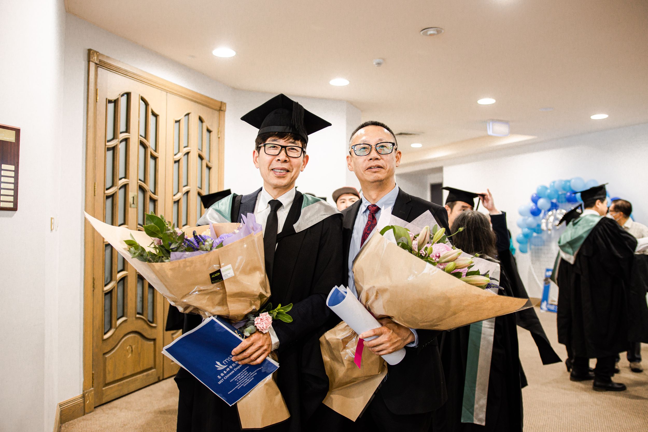 Graduates holding bouquets and diplomas in hallway after ceremony.