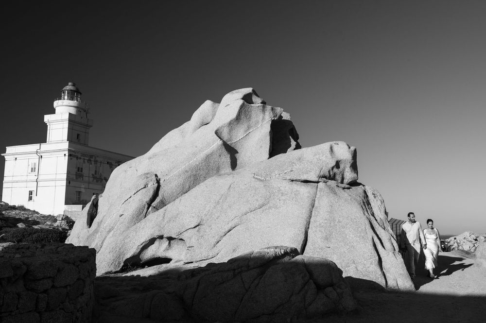 Black and white photo of dramatic granite rock formations with a lighthouse in the background under a clear sky.