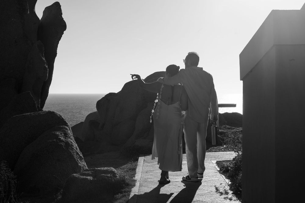 Black and white photo of a couple walking hand in hand toward rocky coastal cliffs under a clear sky.