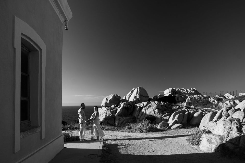 Black and white photo of two people standing near rocky landscape beside a white building under clear sky.