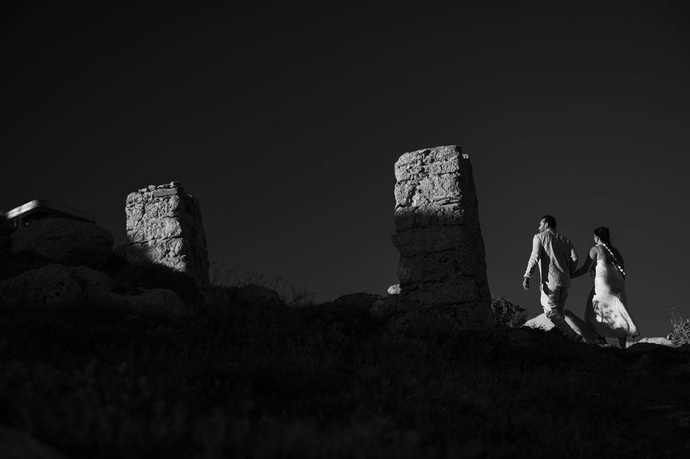 Black and white photo of a couple holding hands walking among ancient stone ruins under a dark sky.