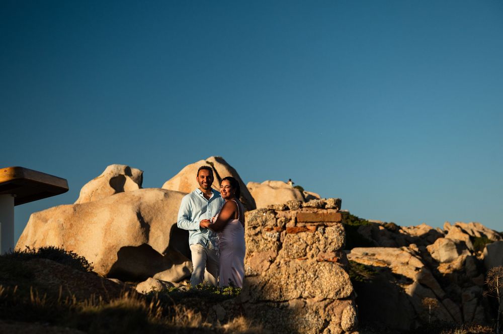 Couple posing together on rocky coastal landscape at golden hour with clear blue sky backdrop.