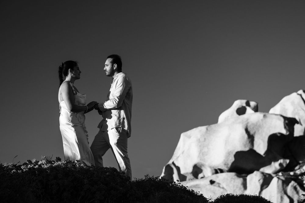 Black and white photo of a couple holding hands on a rocky hilltop, exchanging vows during an outdoor wedding ceremony.