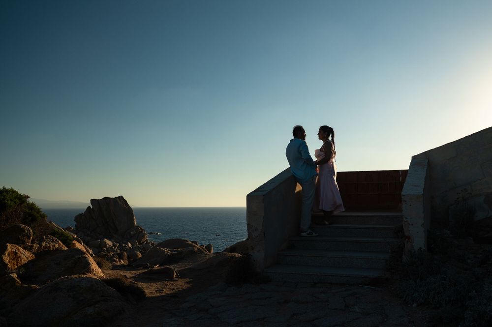 Couple standing on stone steps at sunset overlooking the ocean with rocky coastline in background.