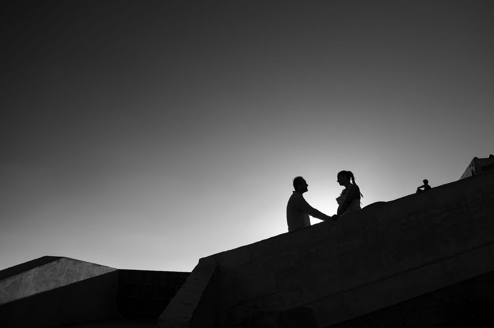 Silhouette of two people facing each other on a rooftop edge against a clear sky in black and white.