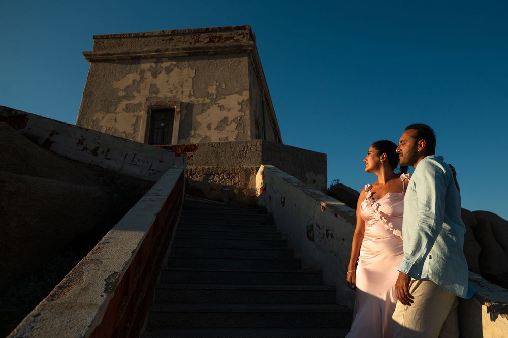 Couple in elegant attire standing on stone steps before a historic building at golden hour.