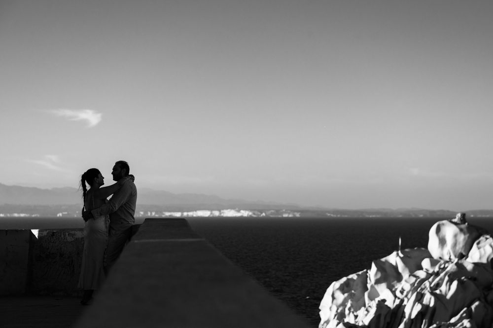 Black and white photo of a couple embracing on a seaside overlook at dusk with city lights in the distance.