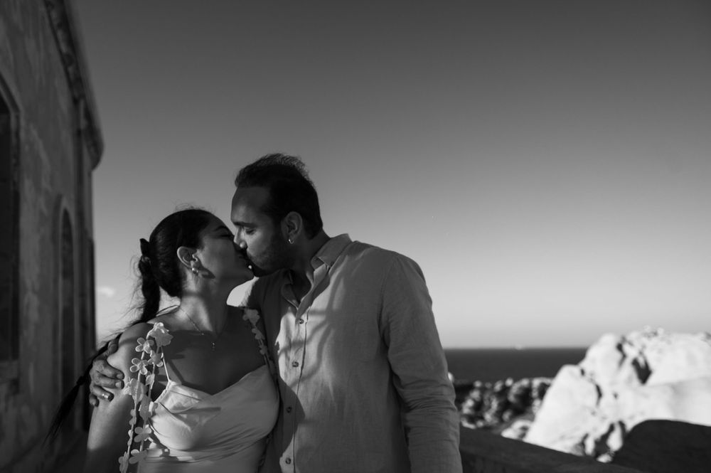 Black and white photo of a couple sharing a romantic kiss outdoors with ocean views in the background.