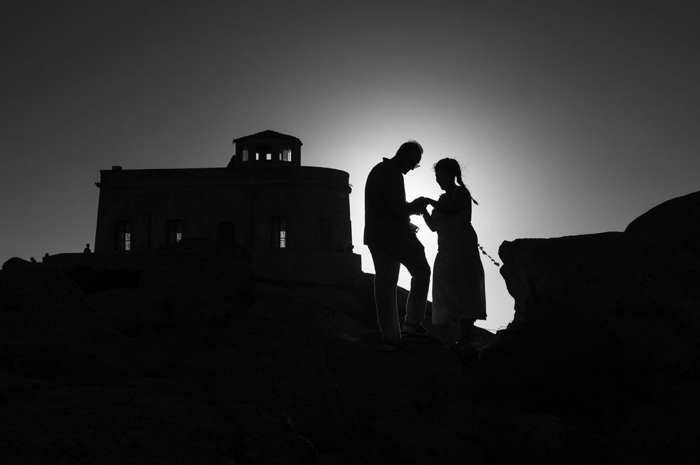 Silhouette of a couple holding hands near an ancient temple ruins against a bright sky in black and white.