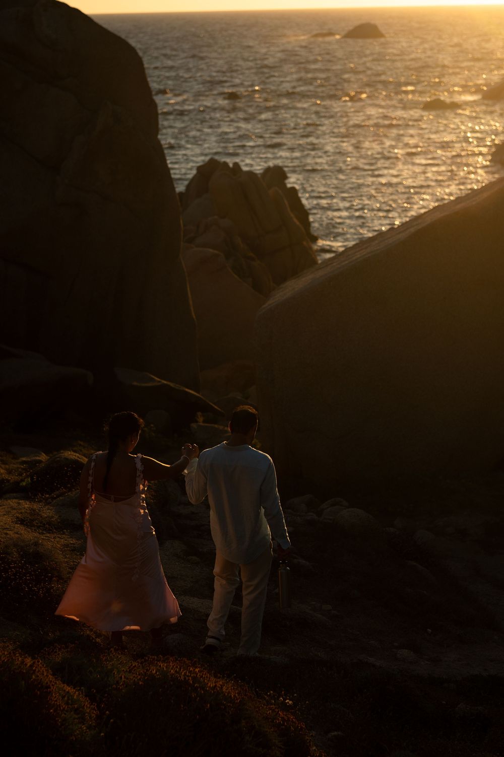 Couple holding hands on rocky coastal cliffs at golden sunset with ocean waves in the background.