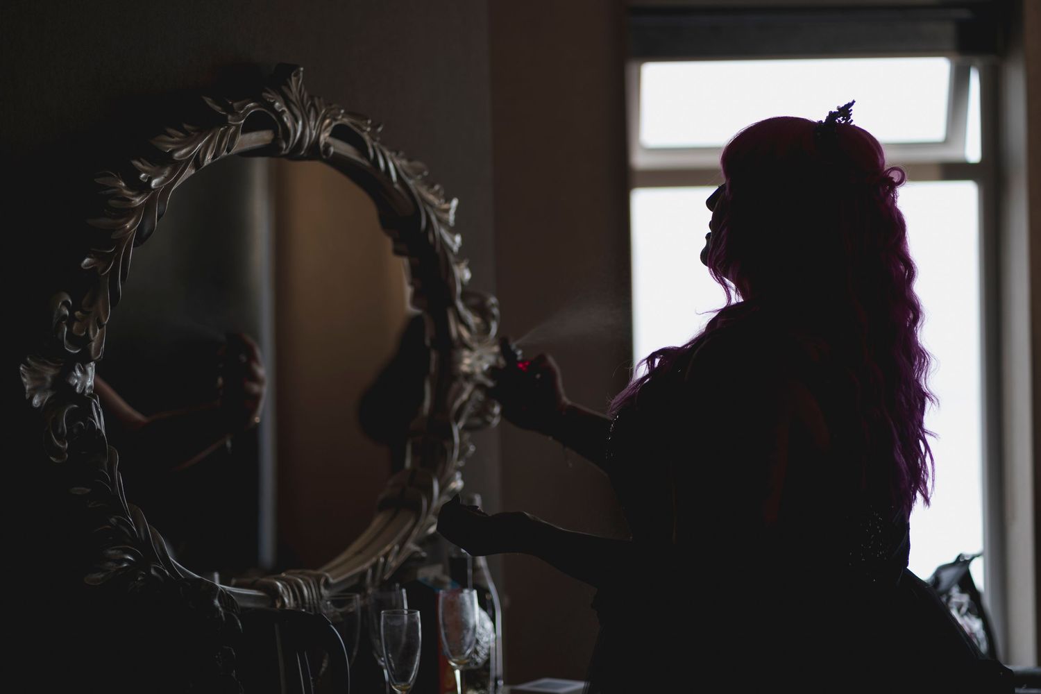 A silhouetted figure stands before a decorated vintage mirror in a darkened room.