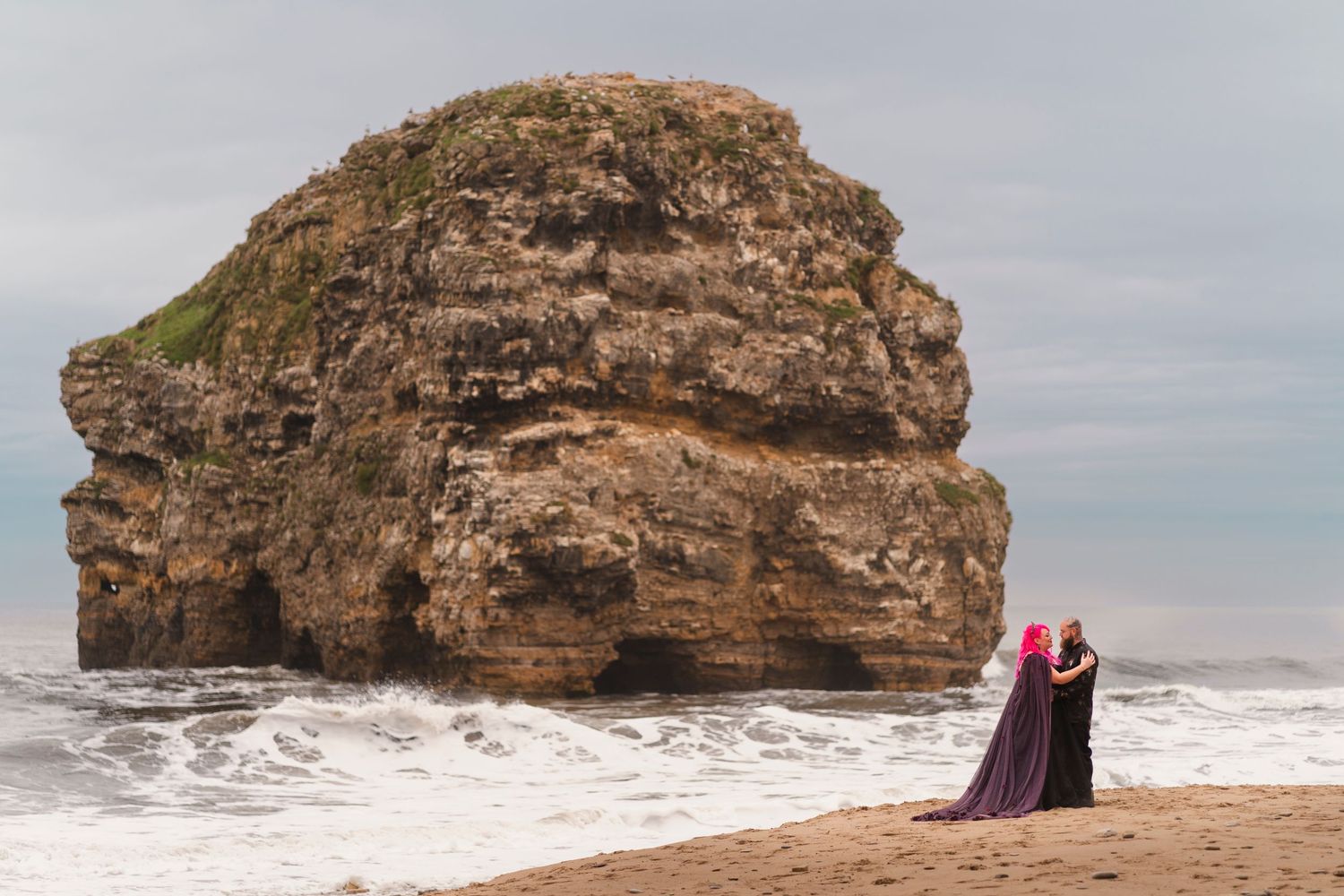 Two figures stand against a dramatic coastal rock formation as waves crash on the shore.