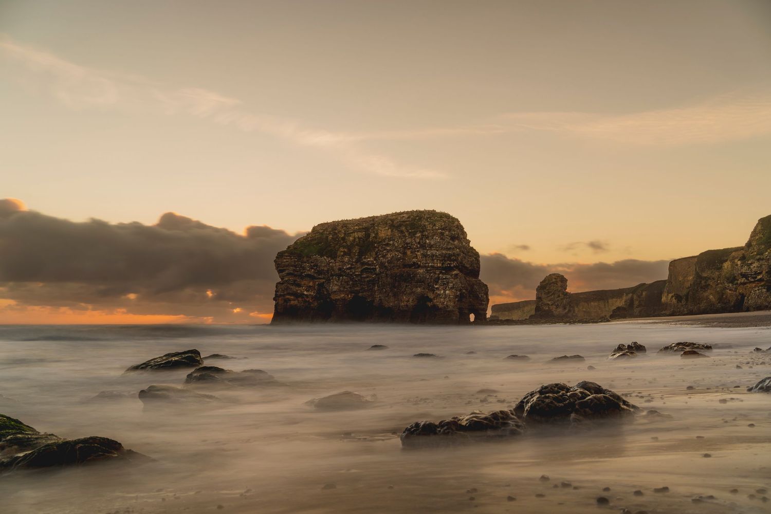 Dramatic sunset view of a rocky coastal beach with large sea stacks and misty waves crashing on the shore.