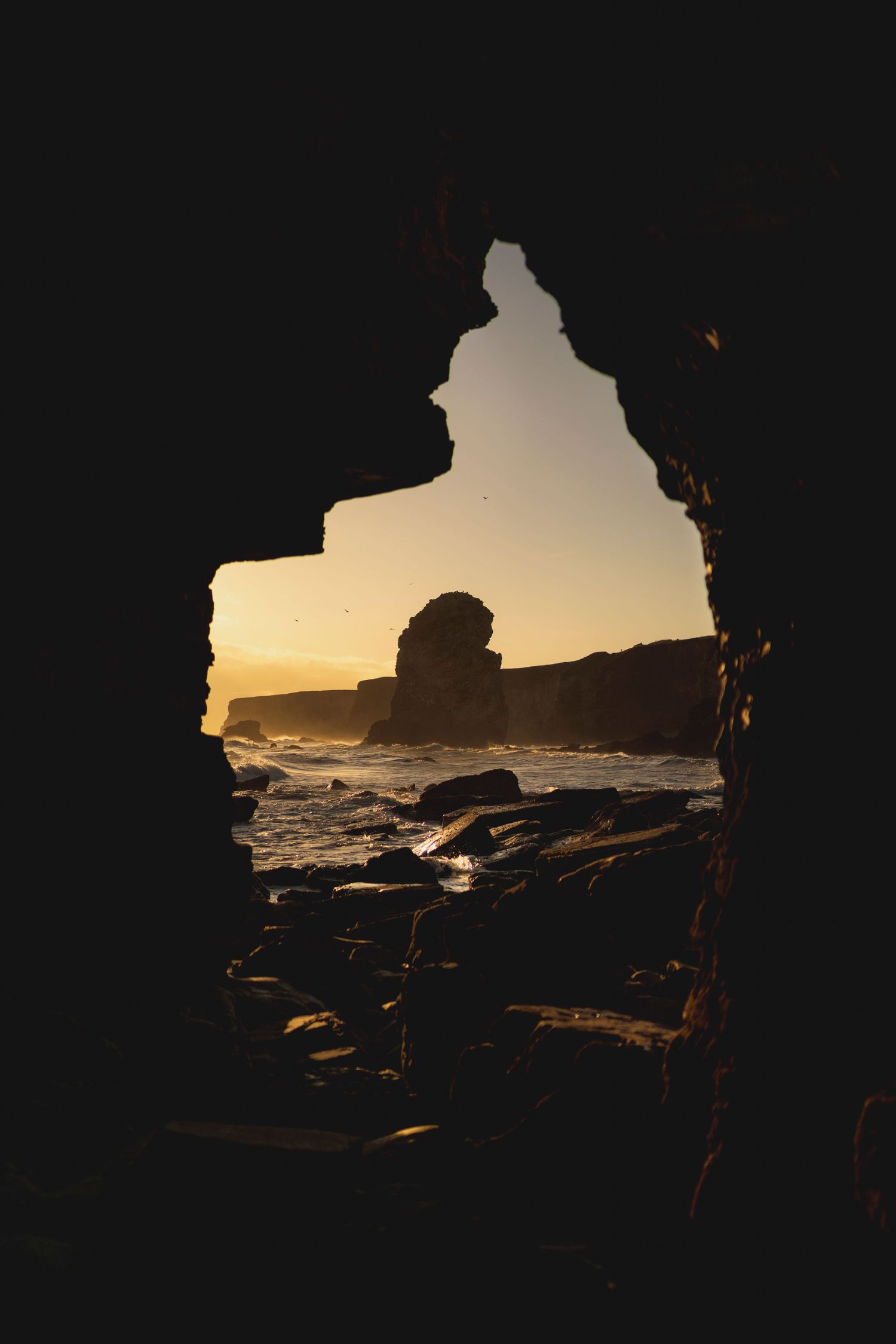Silhouette of coastal rocks and ocean waves viewed through a natural cave opening at golden hour.