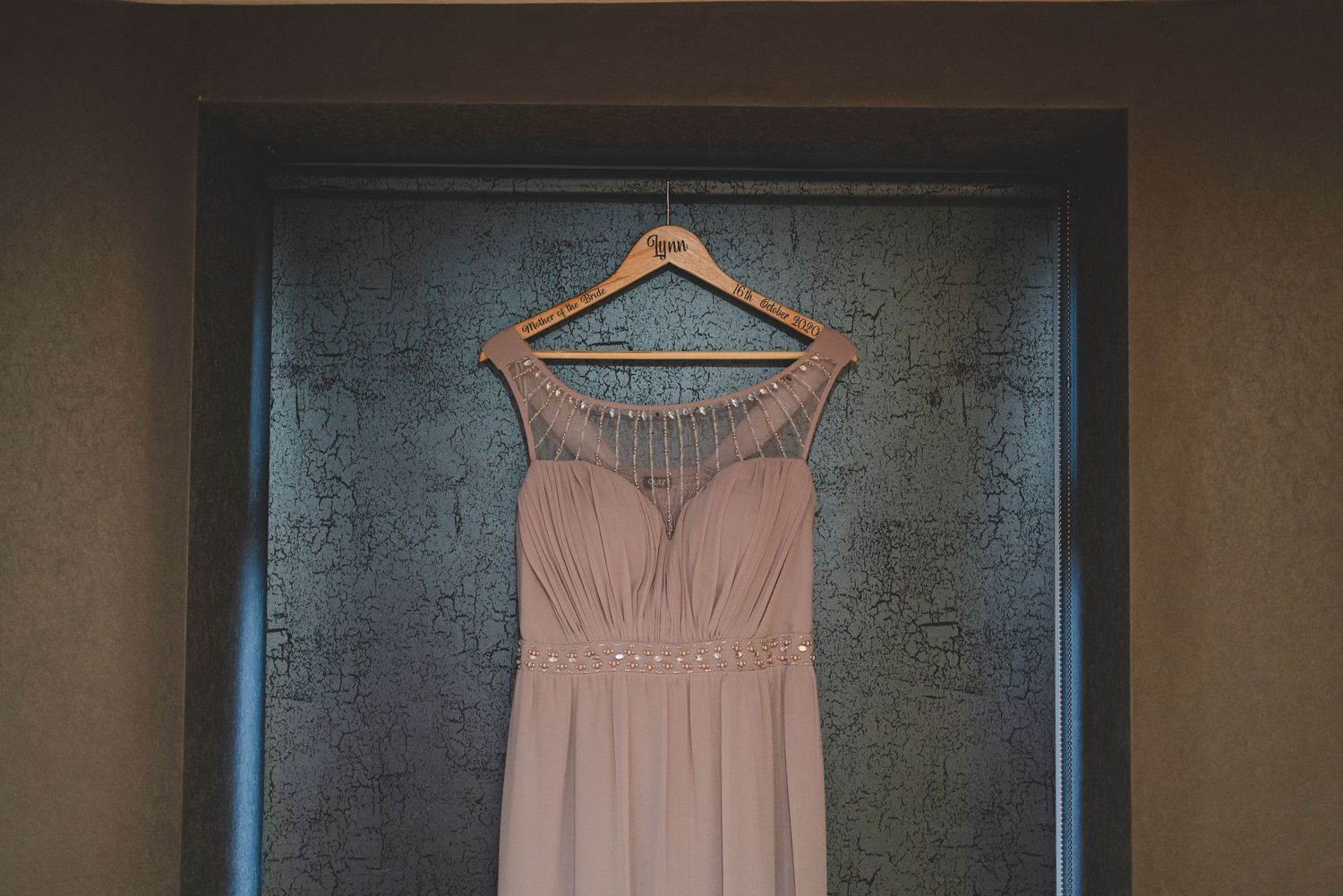 Blush pink bridesmaid dress hanging on wooden hanger against textured grey wall.