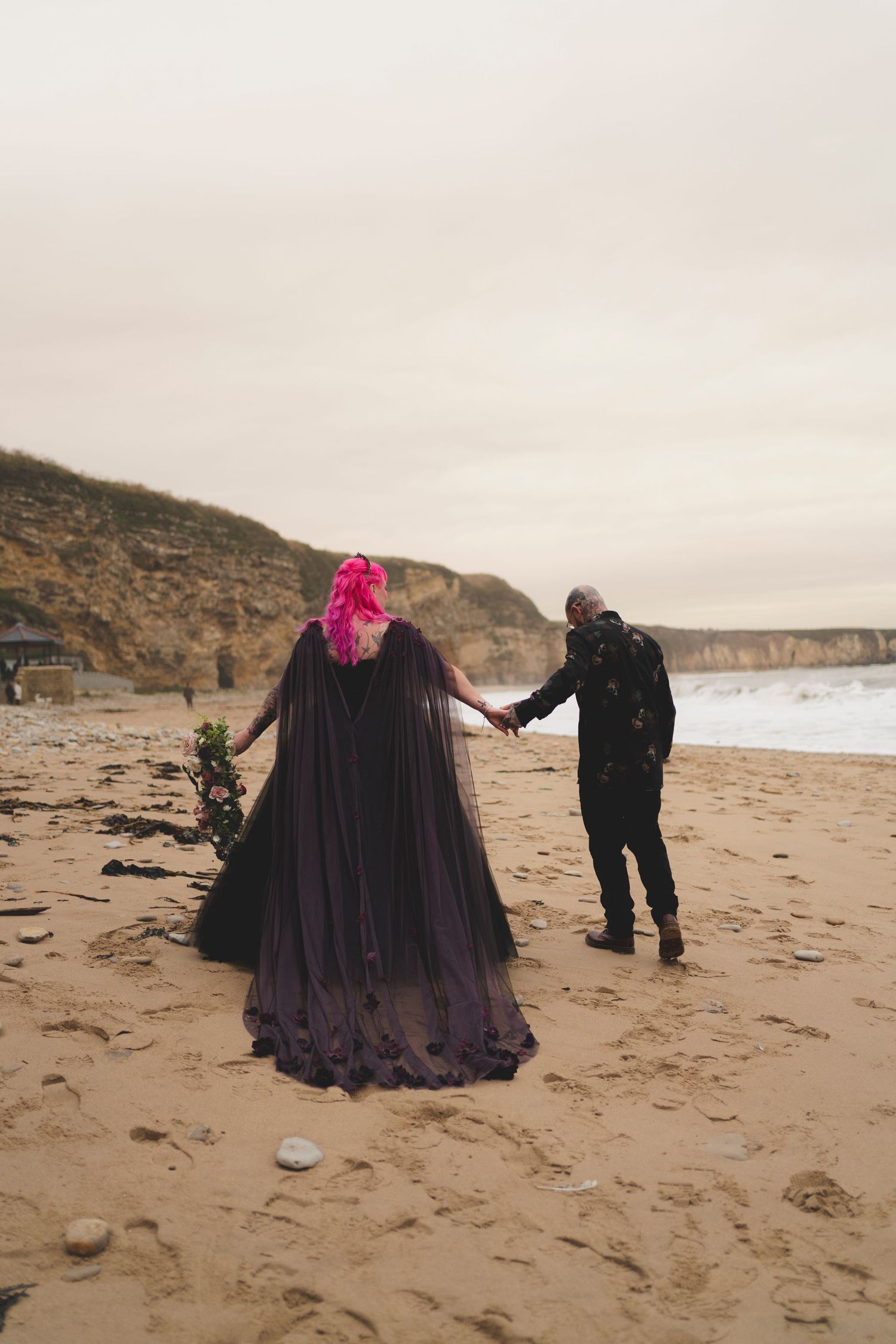 A couple holds hands while walking on a beach with dramatic coastal cliffs.