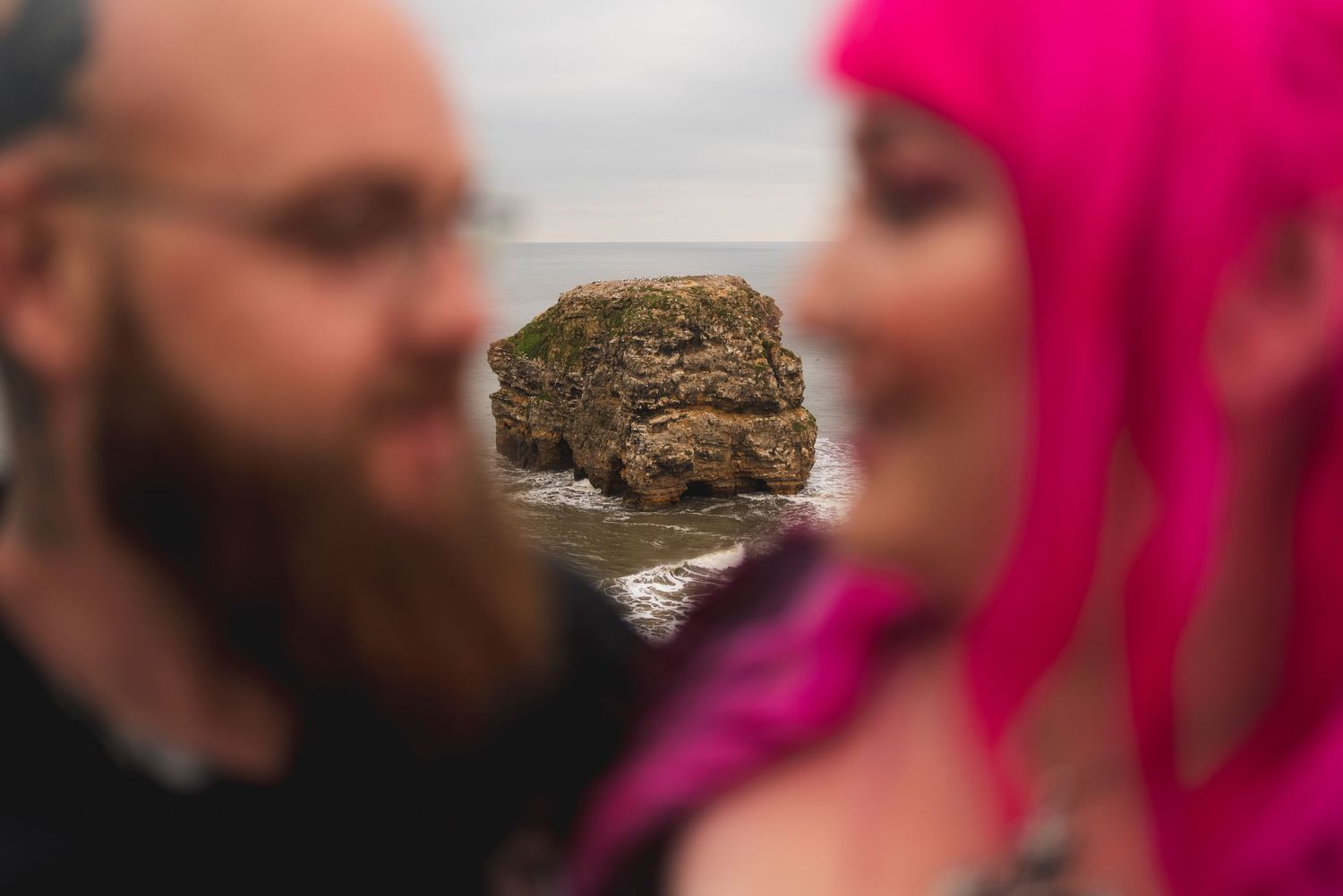 A rock formation in the sea appears blurred in the background of a romantic coastal photoshoot.