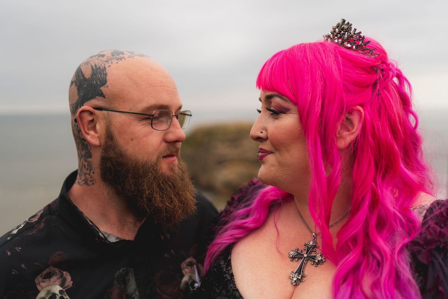 A couple with vibrant pink hair and tattoos share an intimate moment against a moody coastal backdrop.