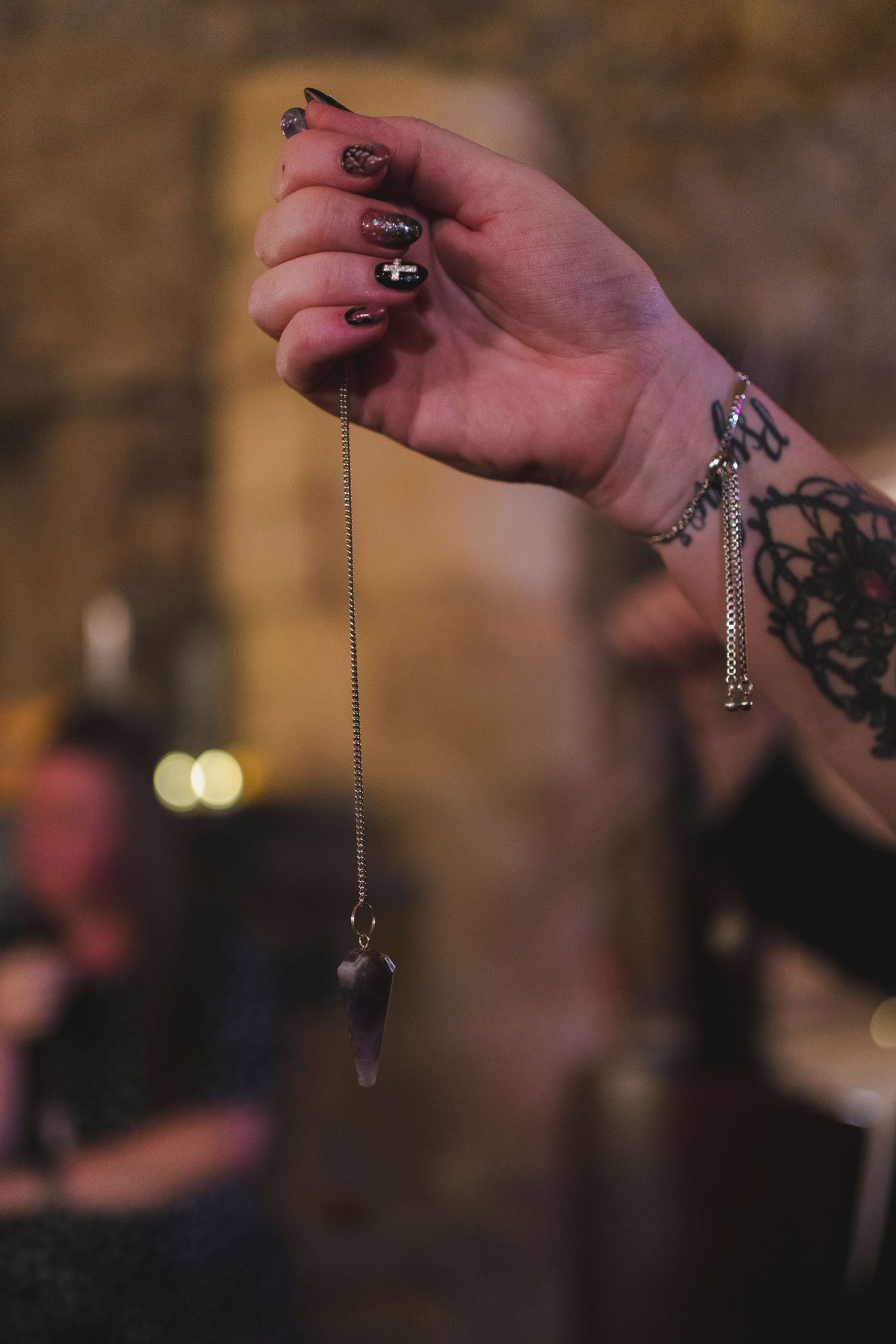 A tattooed hand holds a crystal pendulum against a dark atmospheric background.