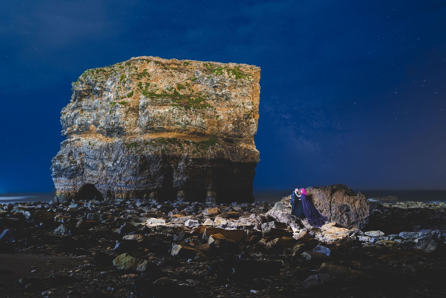 Large limestone sea stack formation illuminated against dark rocky beach at night.
