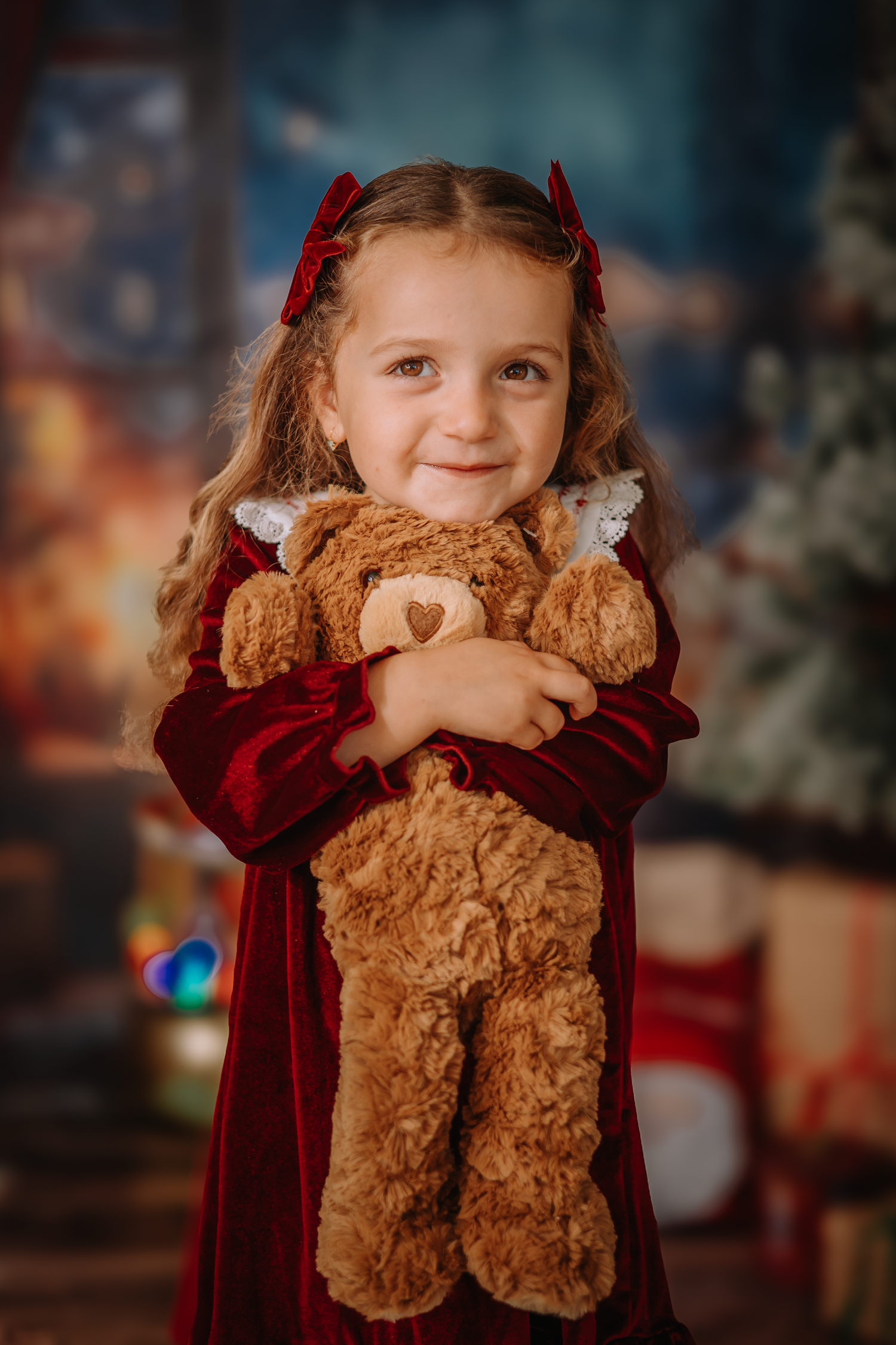 A smiling child wearing a red dress hugs a teddy bear in front of a Christmas backdrop.