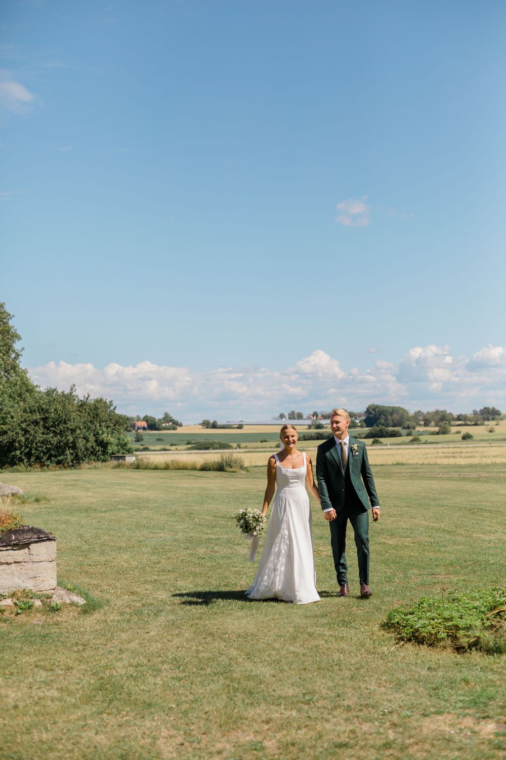 A bride and groom walking together across a sunny field on their wedding day.
