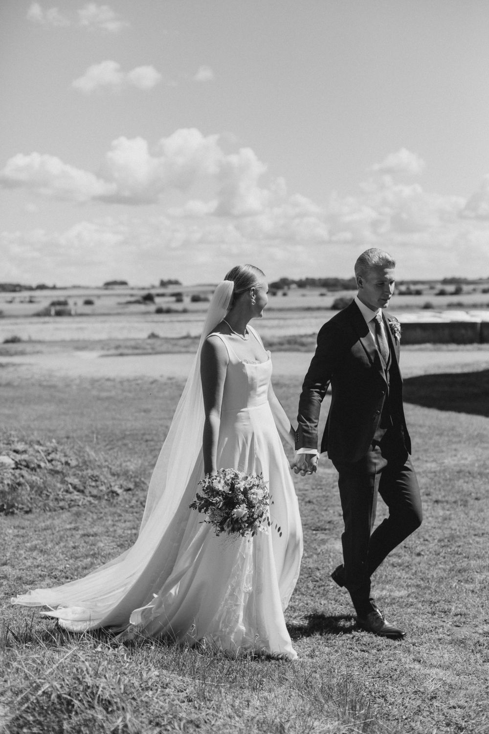 A black and white photo of a couple walking across a field in wedding attire.