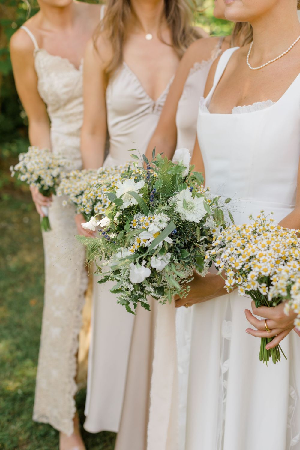 Three bridesmaids in light colored dresses holding delicate white and yellow wildflower bouquets.