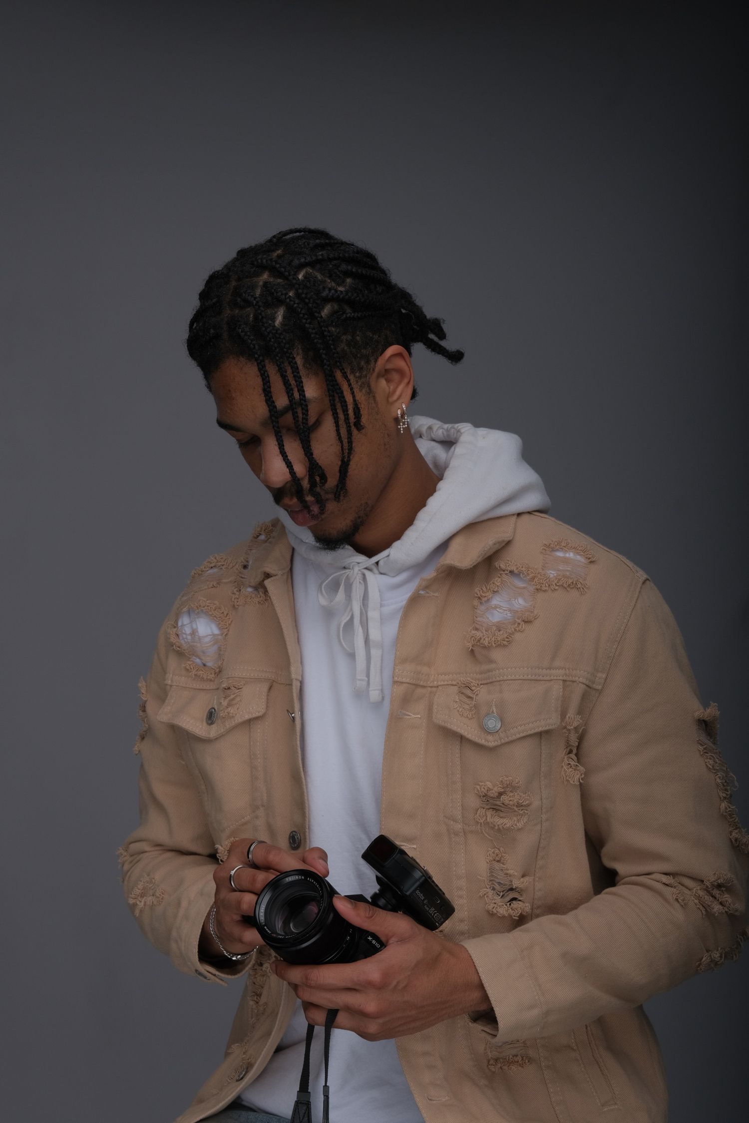 A photographer wearing a distressed tan denim jacket and white hoodie examines their camera against a gray background.