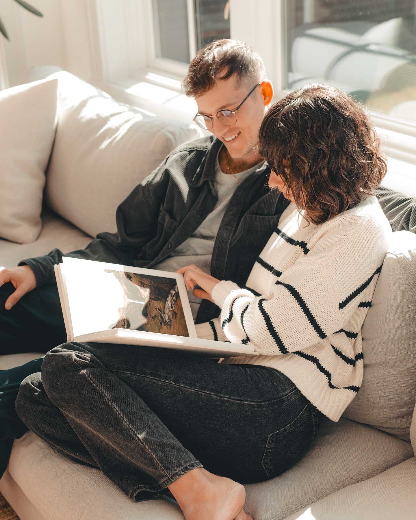 Two people sit close together on a gray couch looking at a photo album in a sunlit room.