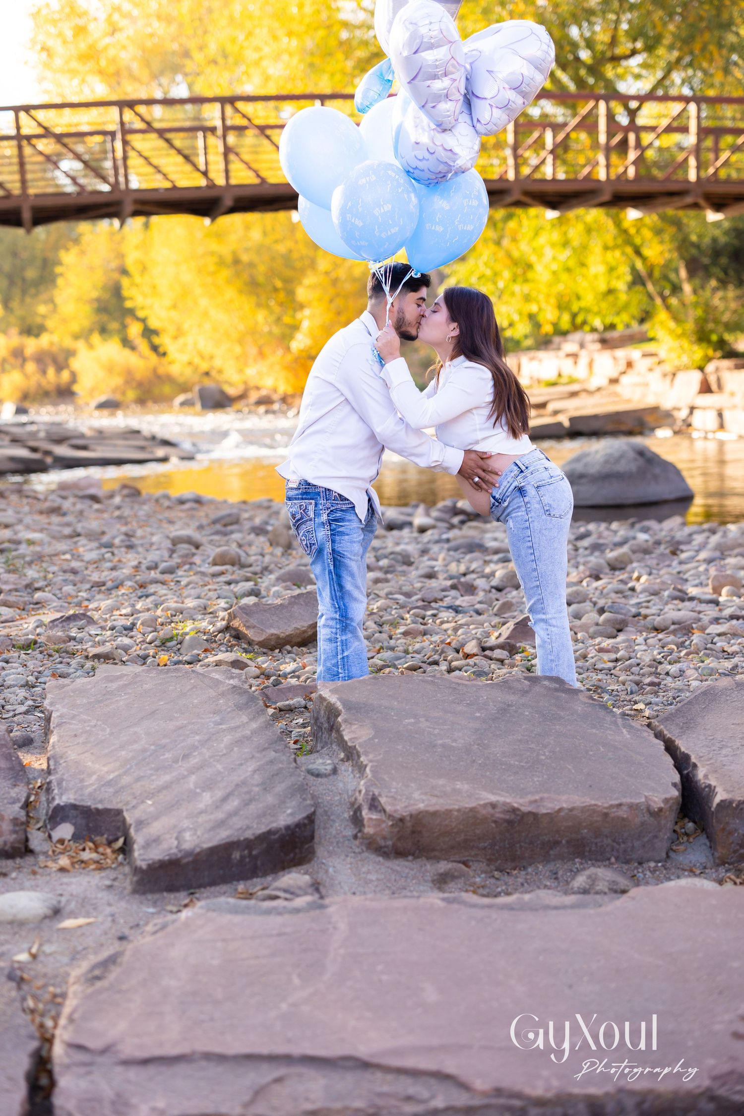 Romantic couple with blue and purple balloons sharing intimate moment on rocky riverside.