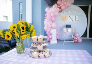 Festive party setup with pink balloon arch, cupcakes, and sunflowers on display table.