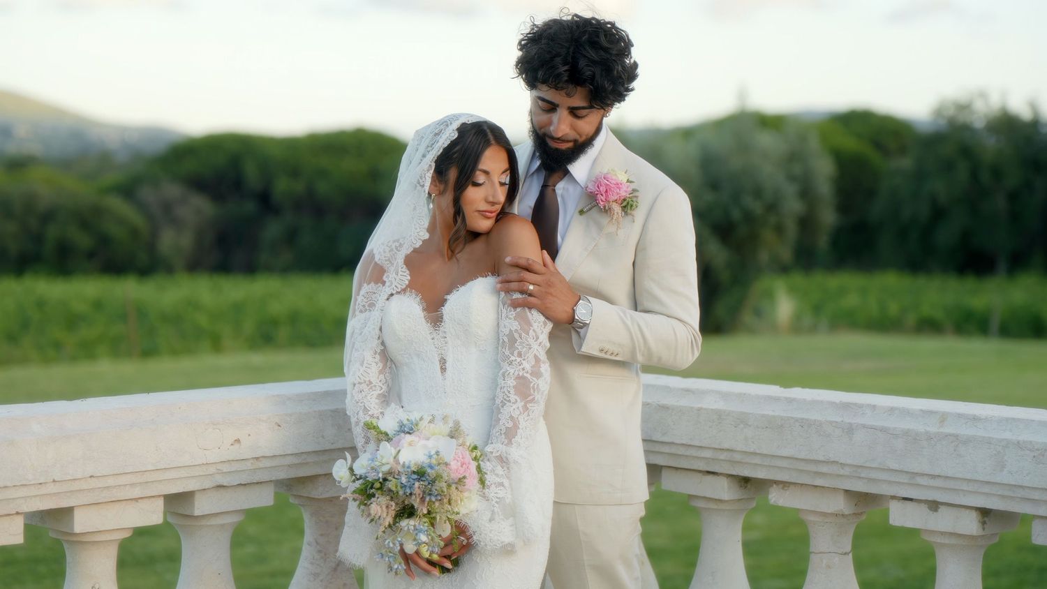 A couple in wedding attire laughs together on a stone balcony overlooking green countryside.