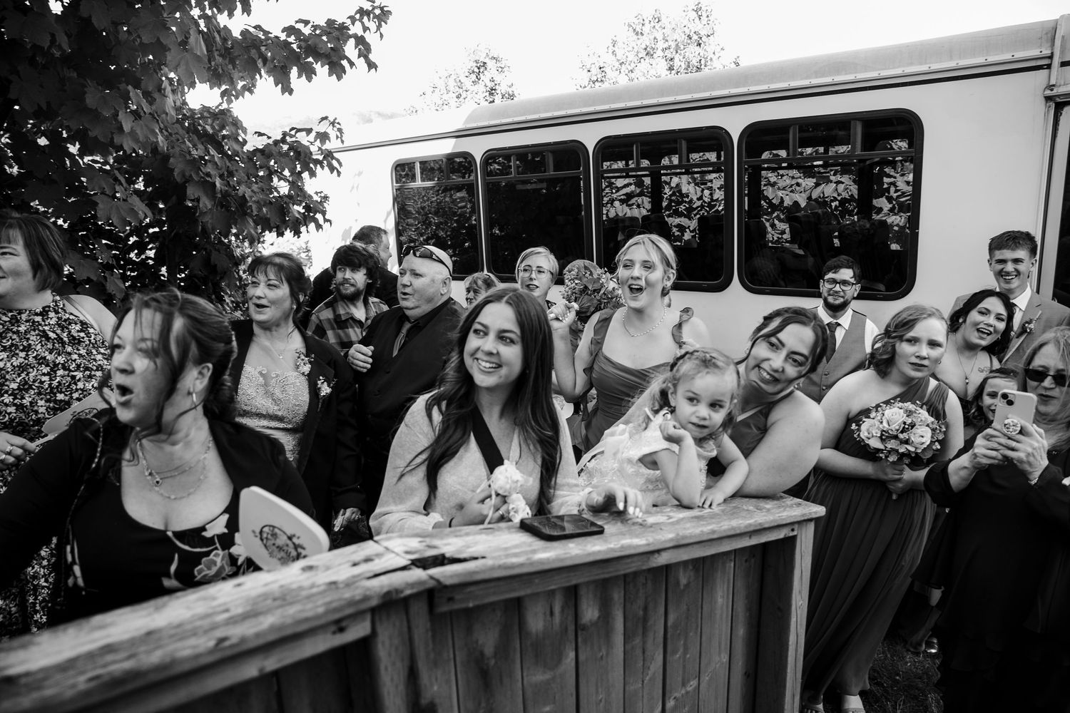 A group of people pose excitedly together against a wooden railing with a bus visible in the background.