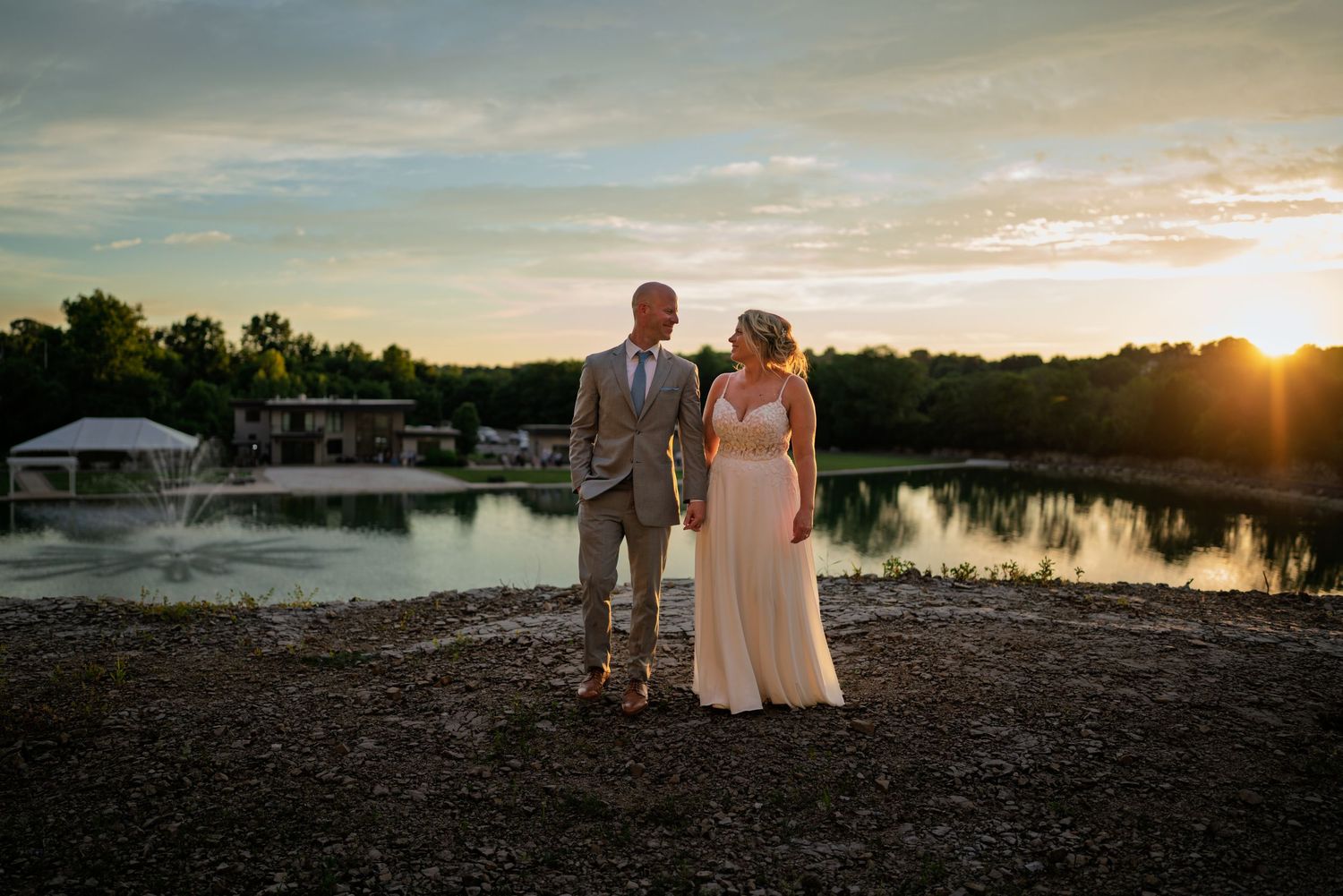 Wedding couple shares an intimate moment by a serene lake with flowing veil and cloudy sky backdrop.