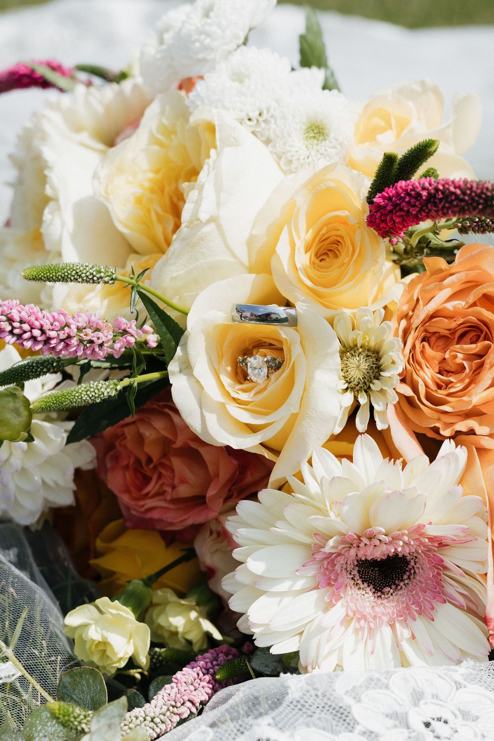 An elegant bridal bouquet featuring peach and cream roses, pink gerbera daisies, and burgundy accents arranged on white lace.