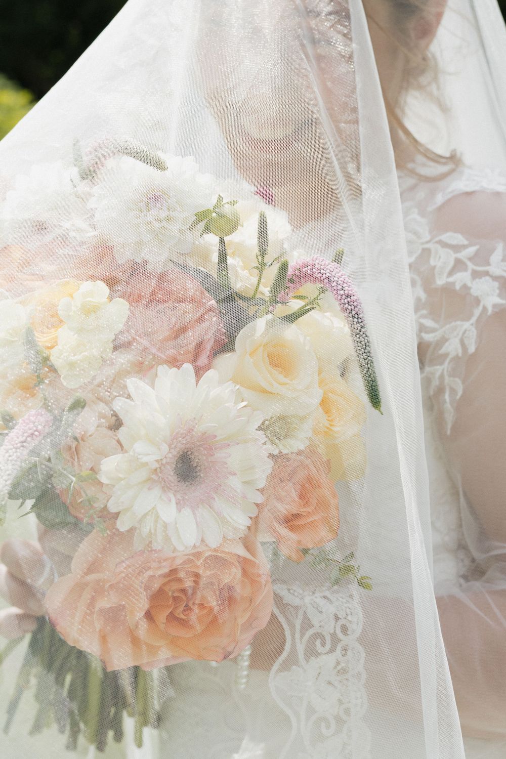 An elegant bridal bouquet of peach roses and white daisies seen through delicate veil fabric.