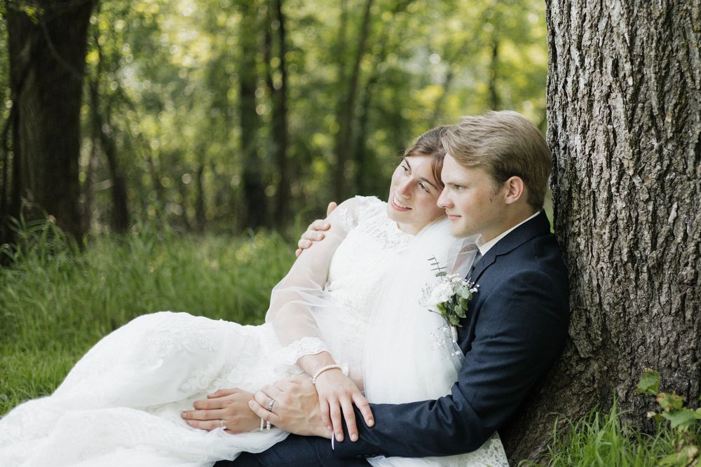 A newlywed couple in formal wedding attire sit together against a tree in a serene forest setting.