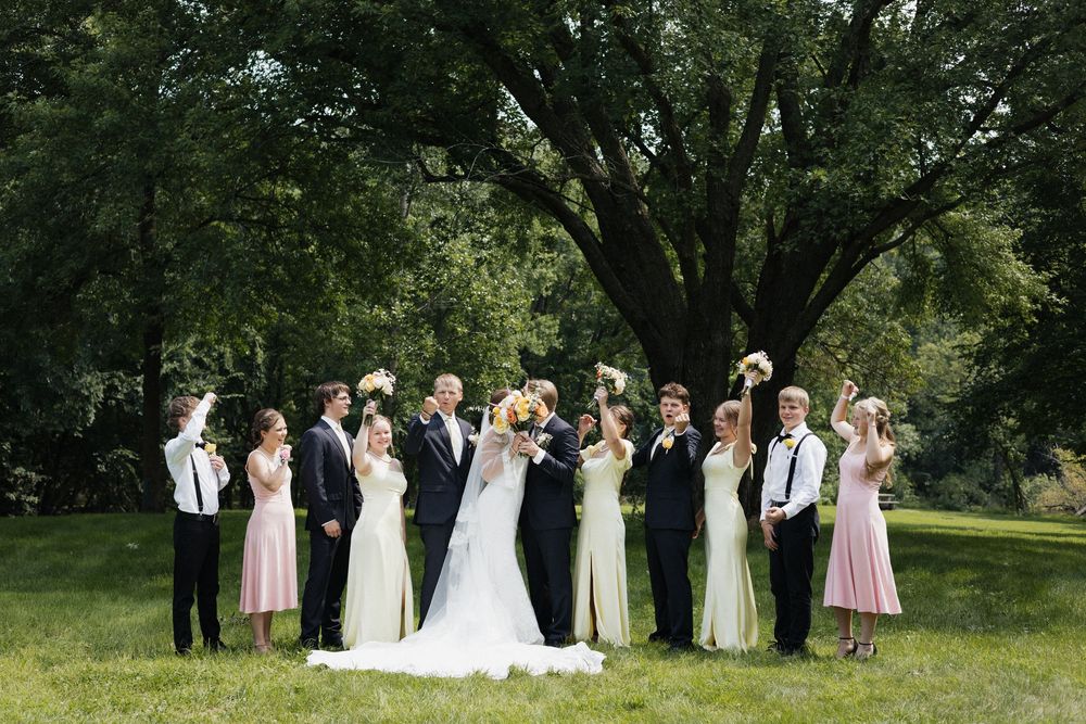 Wedding party poses outdoors under a large tree during a summer celebration.