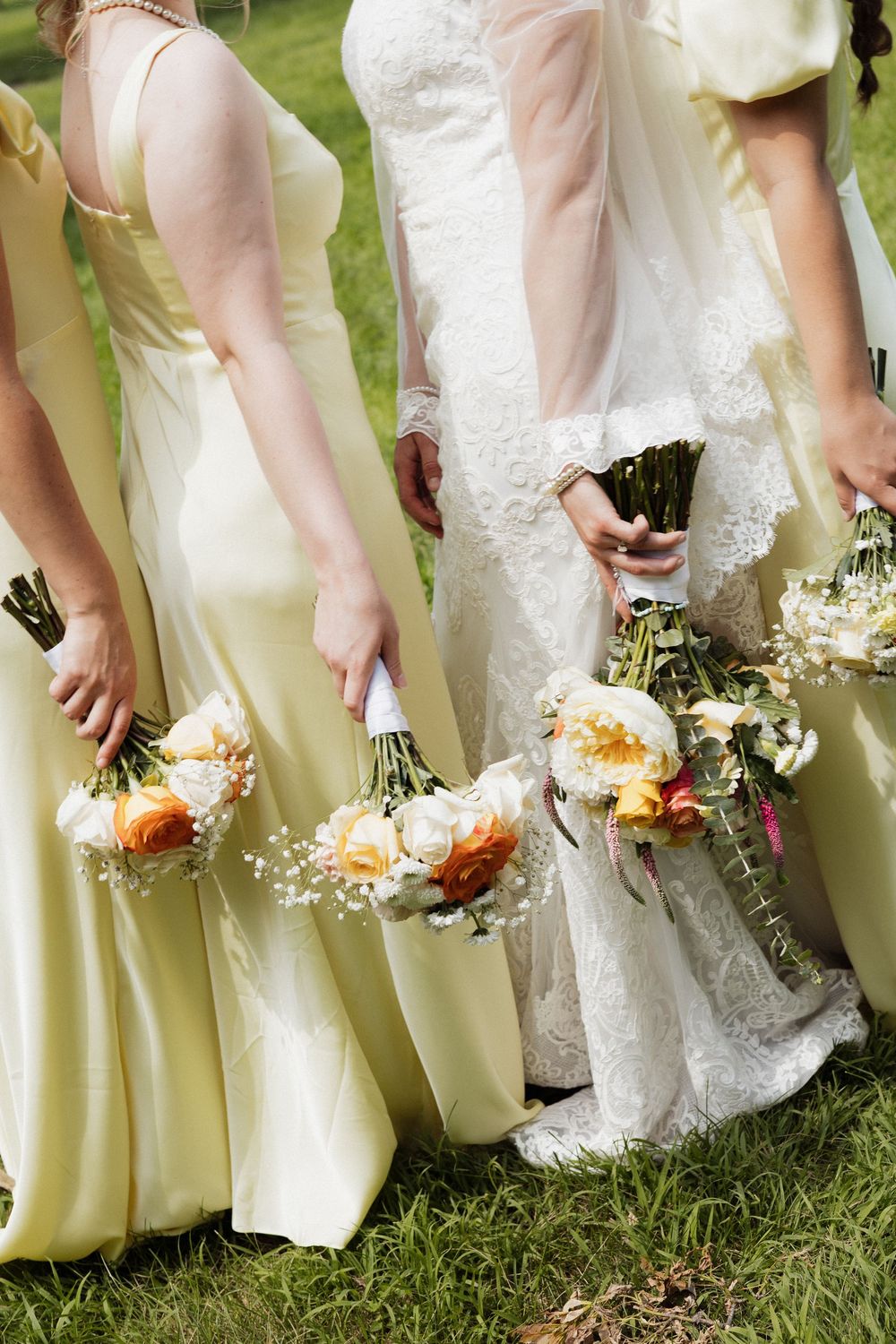 Bridesmaids in pale yellow dresses and bride in white lace gown hold delicate bouquets against green grass.
