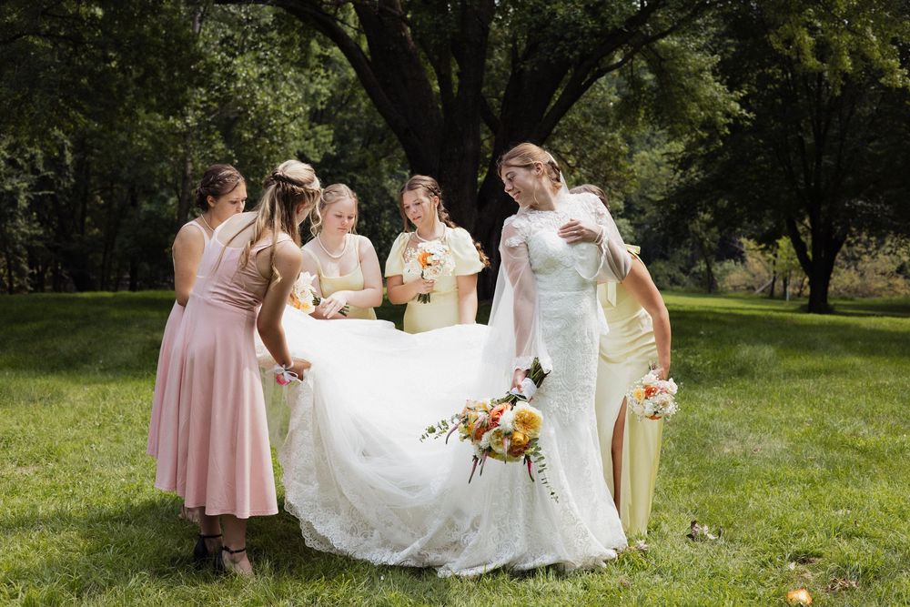 A group in formal attire stands outdoors helping adjust a white wedding dress on a sunny day.