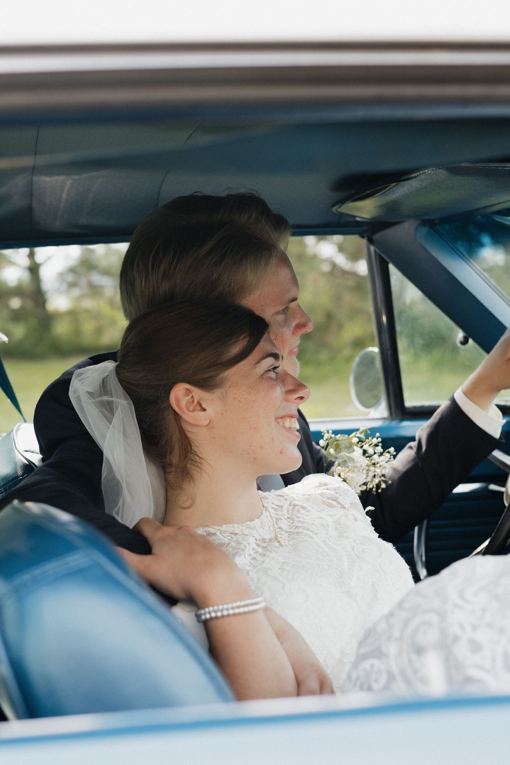 A couple sits in a vintage car on their wedding day with sunlight streaming through the windows.